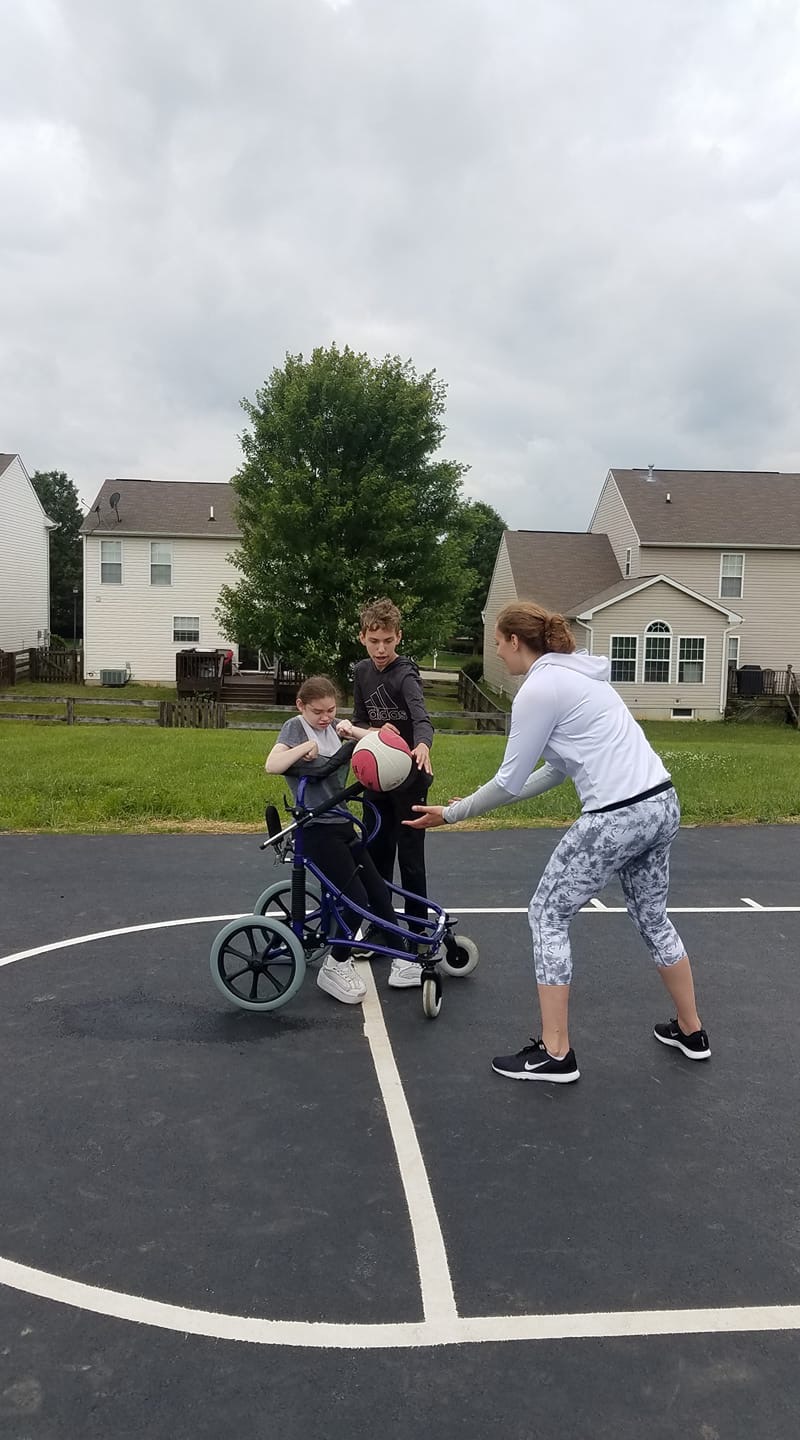 A woman in white playing basketball with her step brother and little sister in a walker.