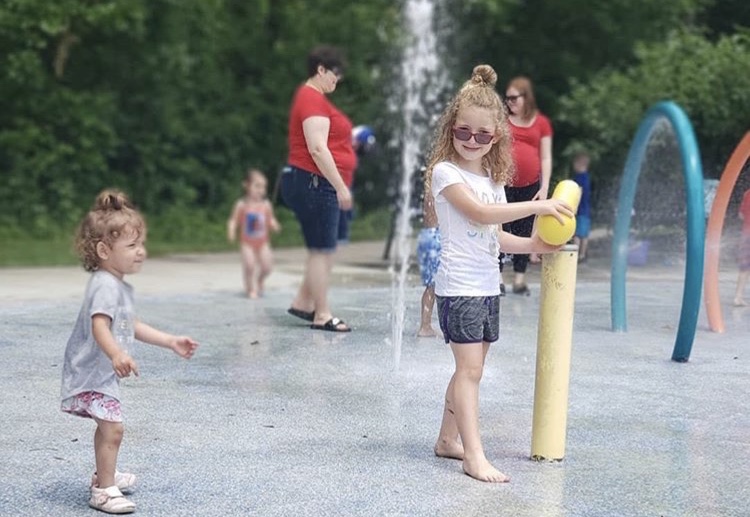 A little girl in a white t shirt and shorts, wearing glasses while playing on a water splash pad. Another little girl is following in her footsteps trying to play with her.